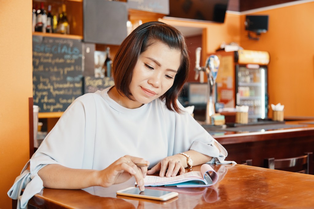 Female bar owner checking phone and writing thoughts and ideas for her business in notepad