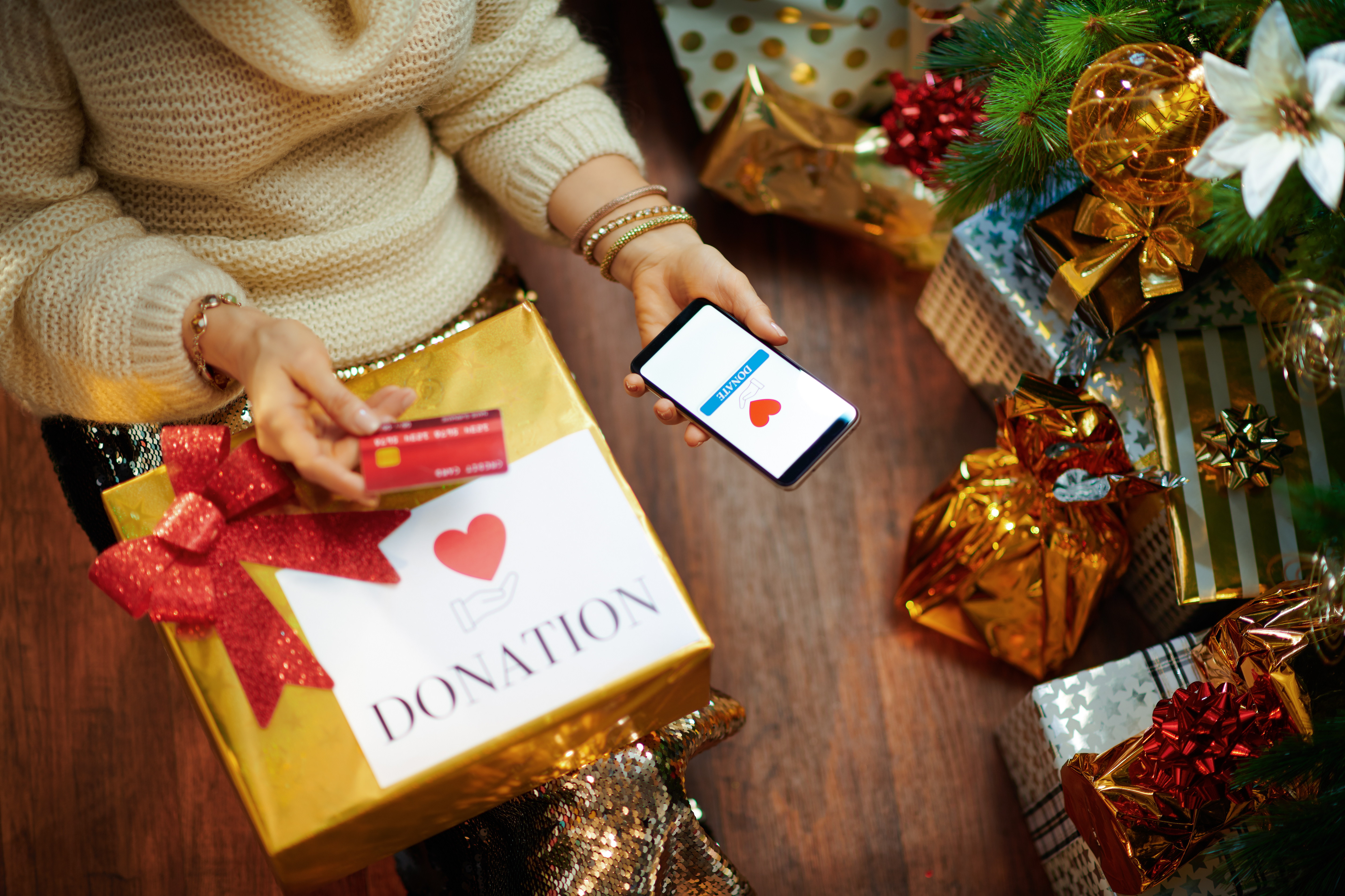 woman in gold sequin skirt and white sweater with credit card making donation via smartphone application under decorated Christmas tree near present boxes.