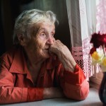 Elderly woman looks sadly out the window.