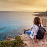 A stylish young woman traveler watches a beautiful sunset on the rocks of a beach